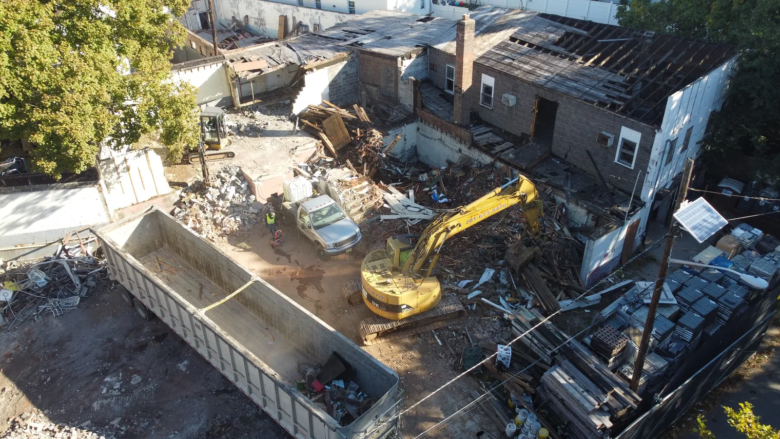 Aerial view of a teardown lot with an excavator loading a debris trailer, a mini excavator and a crew member, demolition contractor