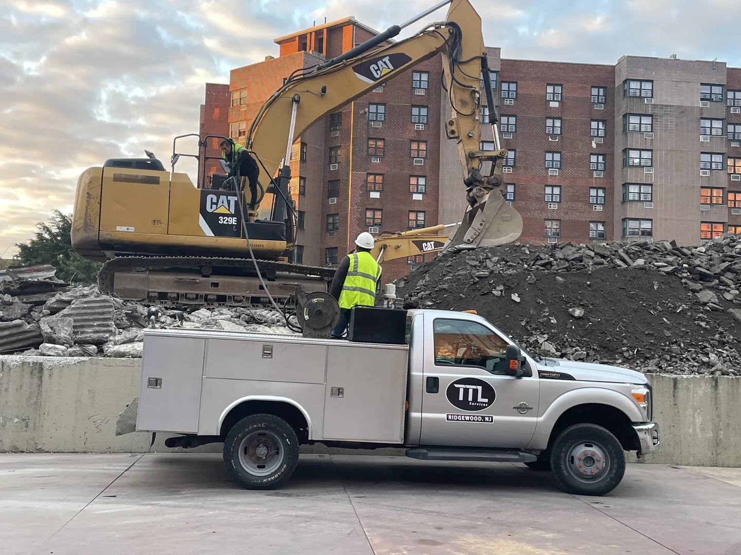 Company service truck parked below an excavator on a demolition pile, a crew member climbing into the cab, apartment buildings behind