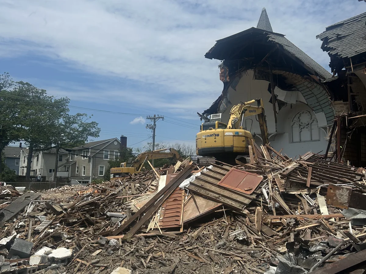 Excavator working at the torn open wall of a church building below its steeple, a large debris pile in front, asbestos abatement and demolition