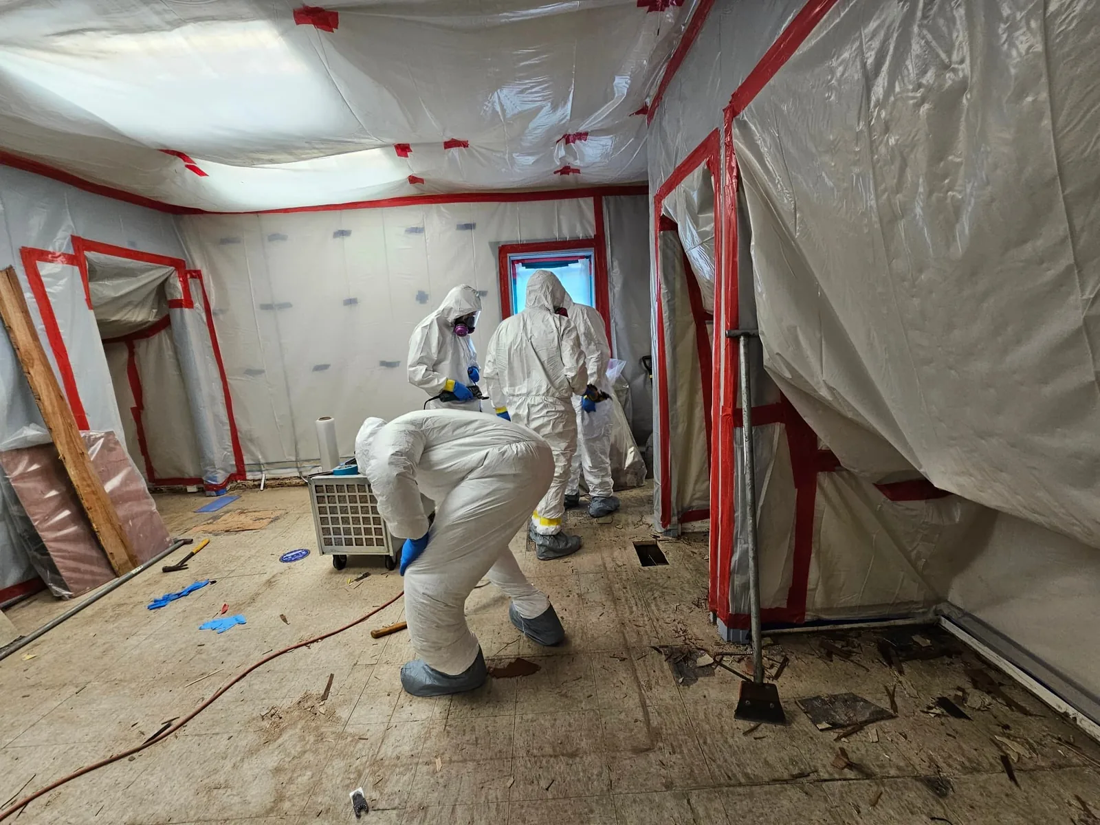 Workers in protective suits and respirators inside a room sealed with plastic sheeting and red tape, old flooring partly removed, asbestos abatement