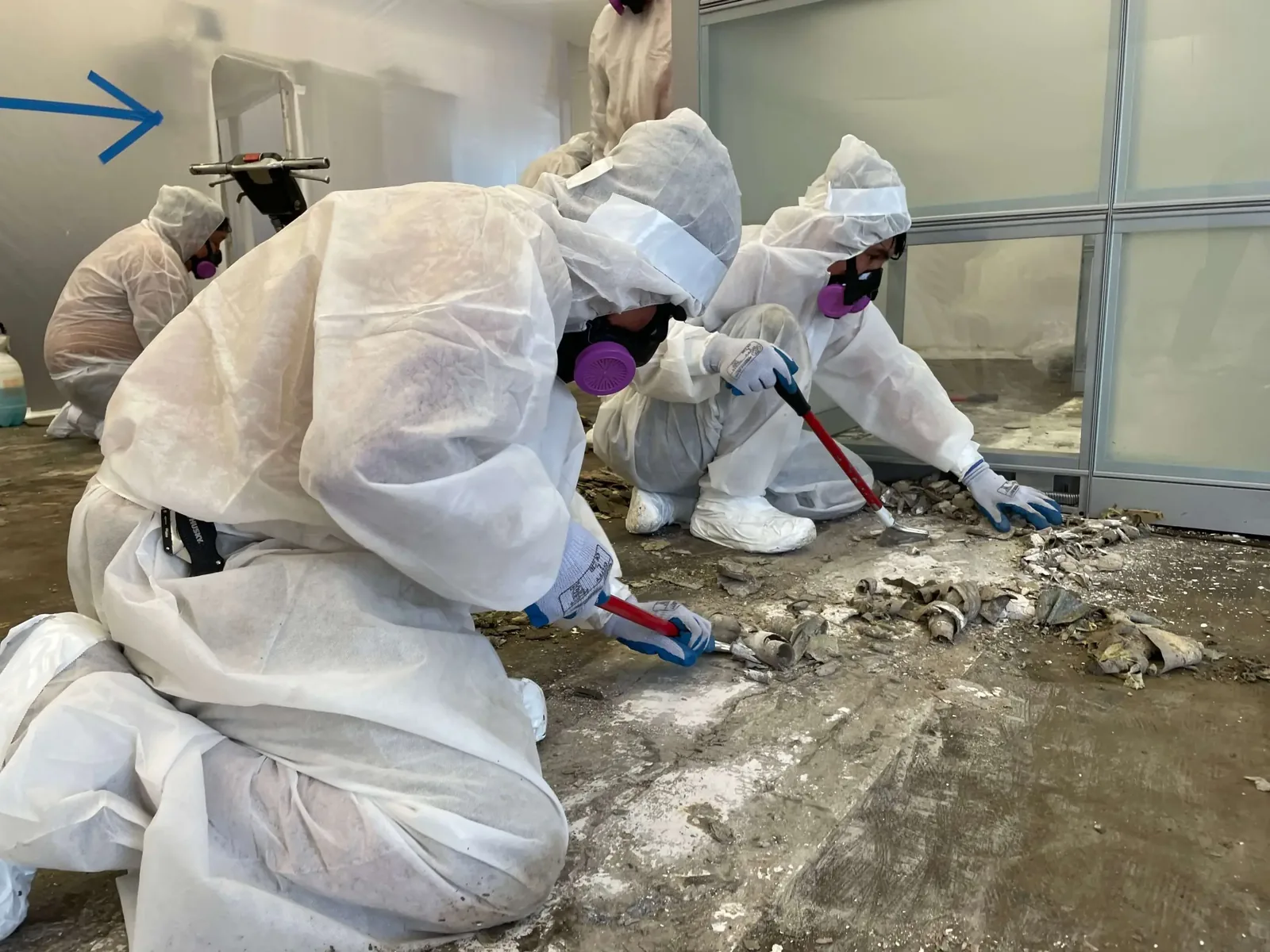 Workers in protective suits and respirators scraping old flooring inside a room sealed with plastic sheeting, asbestos abatement