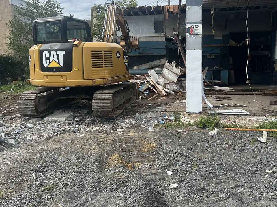Excavator in front of a gutted one story storefront building coming down on a gravel lot, commercial demolition