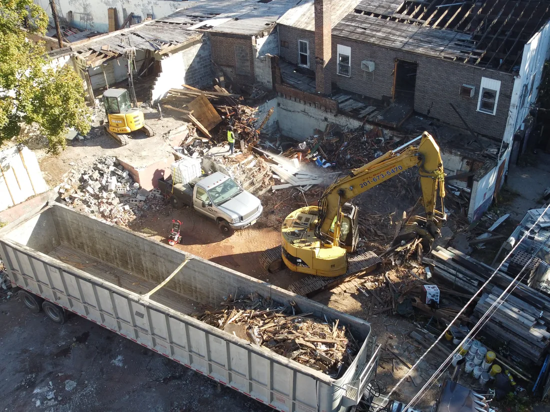 Aerial view of an excavator loading demolition debris into an open top trailer as a worker sprays water to keep the dust down, debris removal
