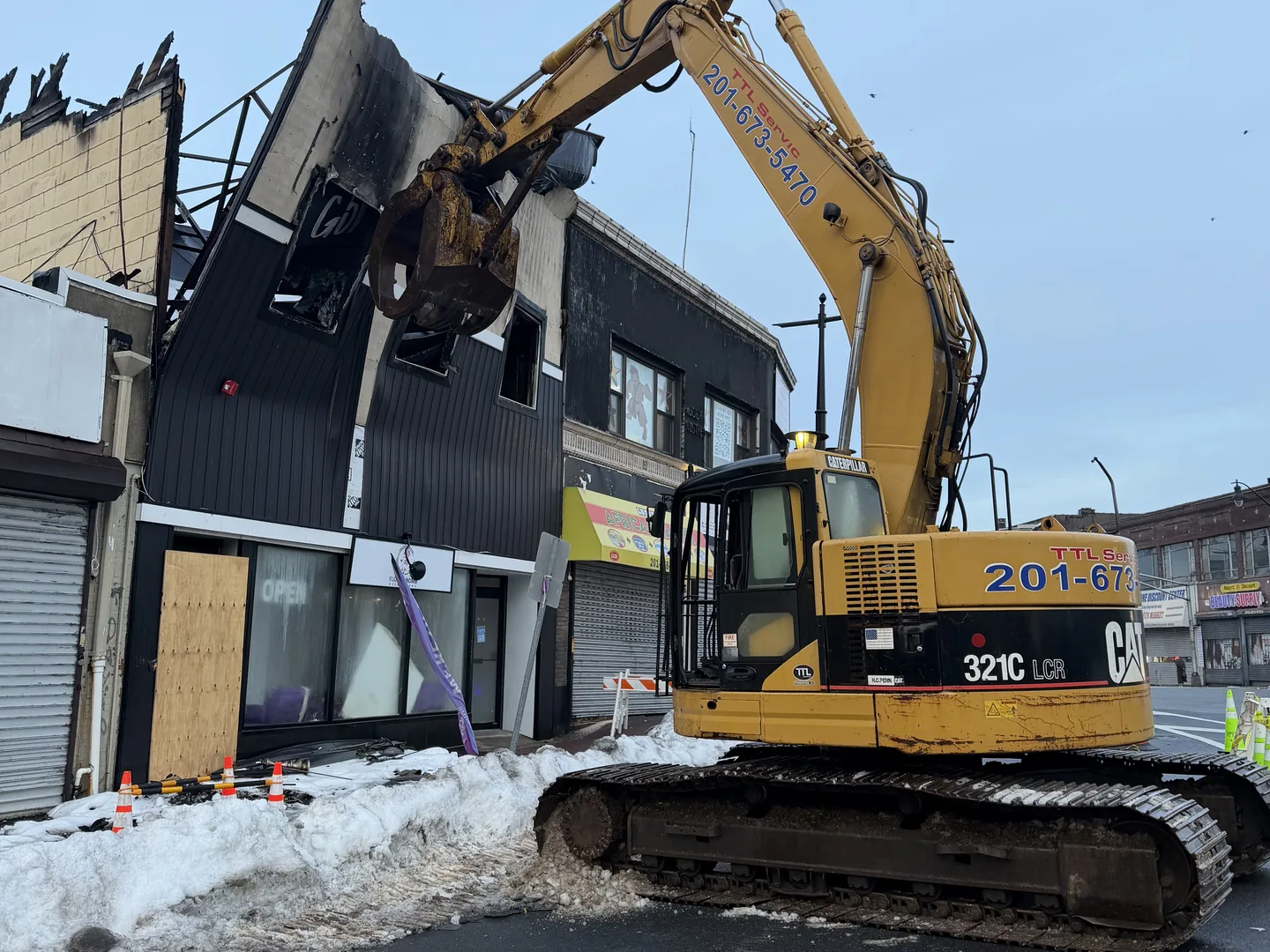 Excavator with a grapple pulling down the burned front wall of a two story storefront building, snow along the curb, emergency demolition