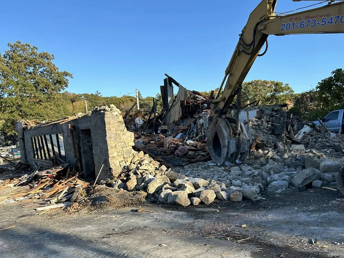 Excavator grapple over the stone walls and burned framing of a fire damaged building, stone rubble in front, emergency demolition