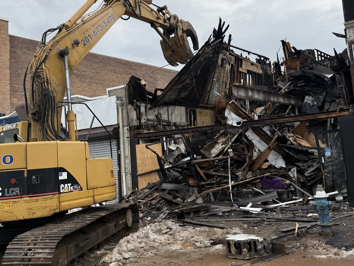 Excavator grapple lifting charred framing from a burned storefront building, a fire hydrant in front, emergency demolition