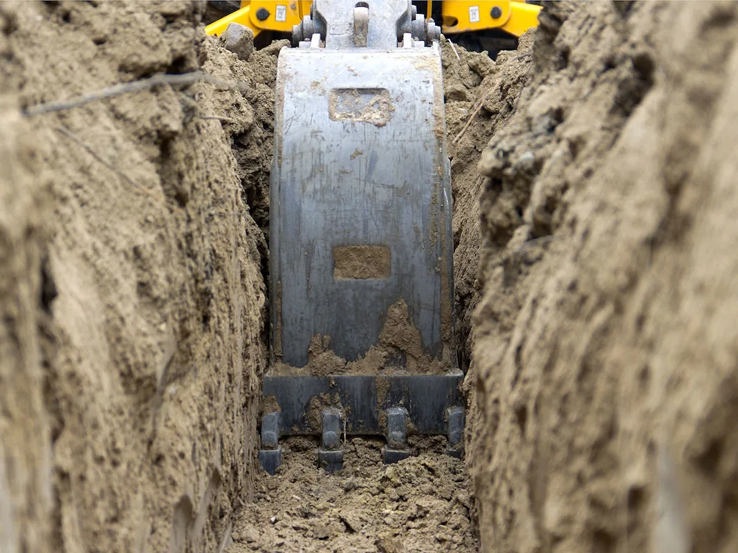 Excavator bucket digging the bottom of a narrow freshly dug trench, excavation