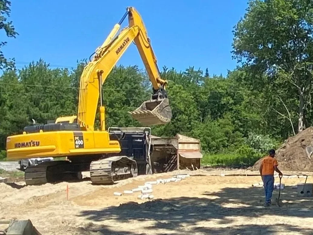 Large tracked excavator loading spoils into a dump truck on a cleared sandy lot as a small crew works by a dirt pile, excavation