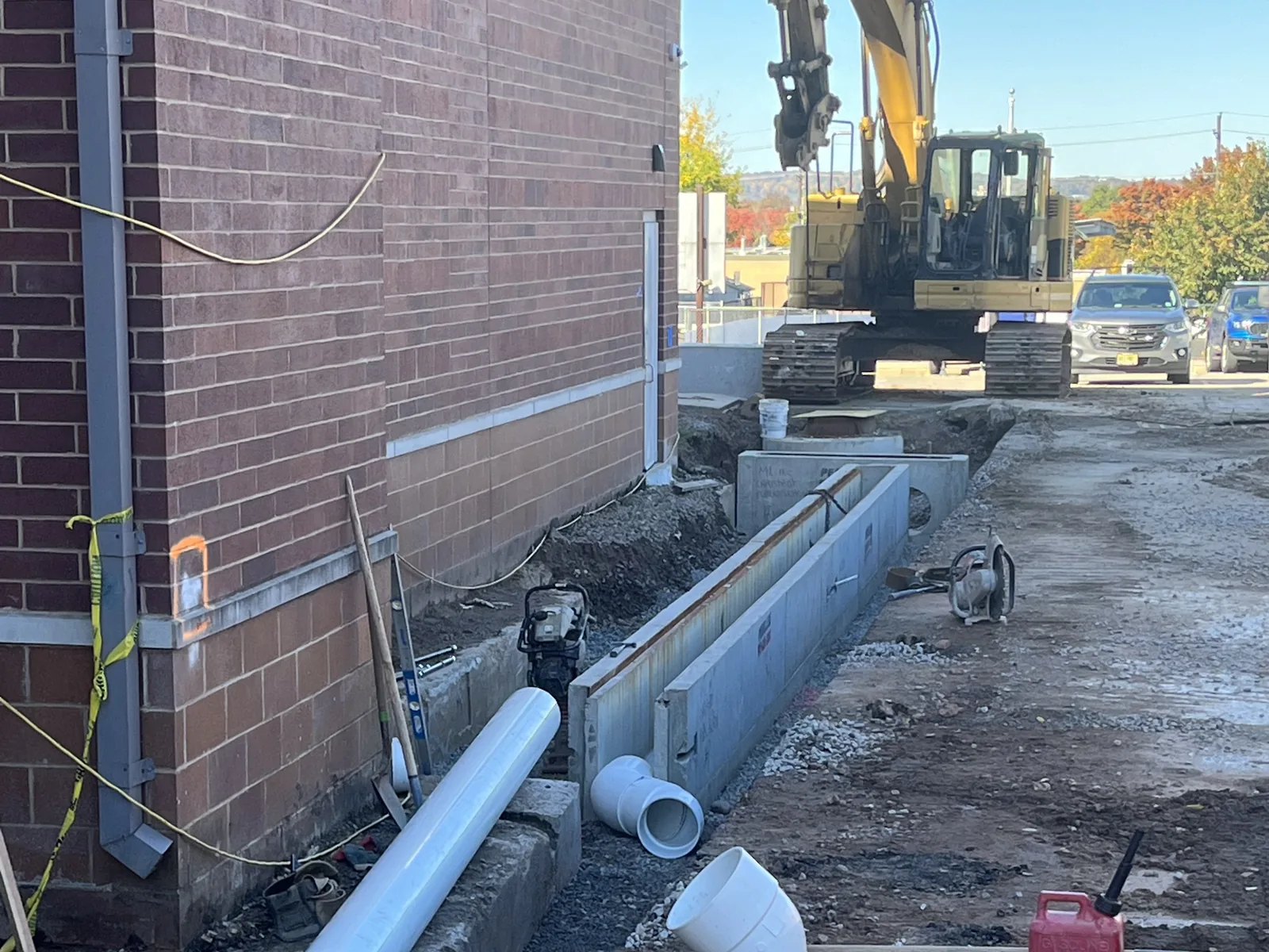 Trench dug along a brick building with drain channel sections and white pipe laid out, an excavator behind, excavation