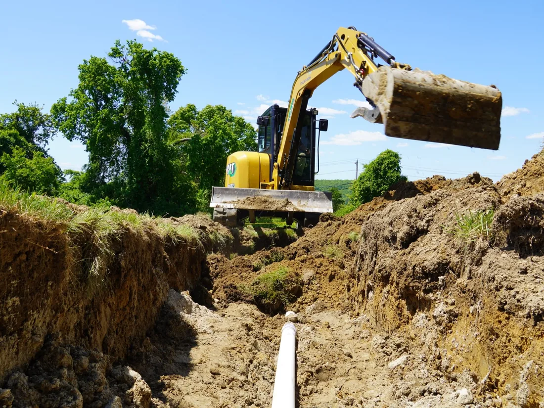 Compact excavator digging a utility trench across a grassy lot, a white pipe along the trench bottom and spoil banks on both sides, excavation
