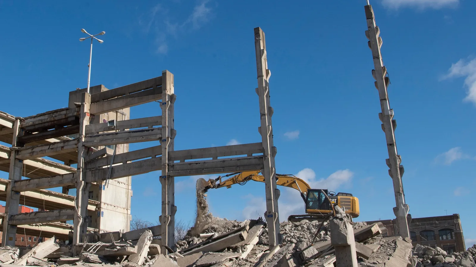Excavator dropping rubble beside the last standing columns of a multi story parking garage coming down, demolition contractor