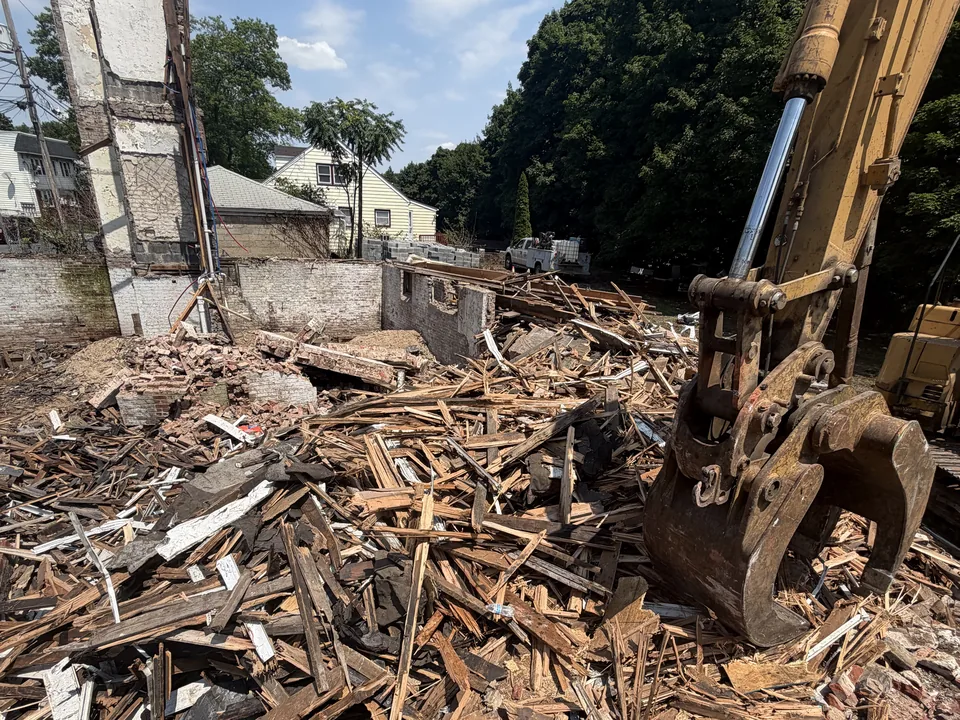 Excavator grapple over the splintered wood debris of a torn down house, its brick chimney still standing, house demolition
