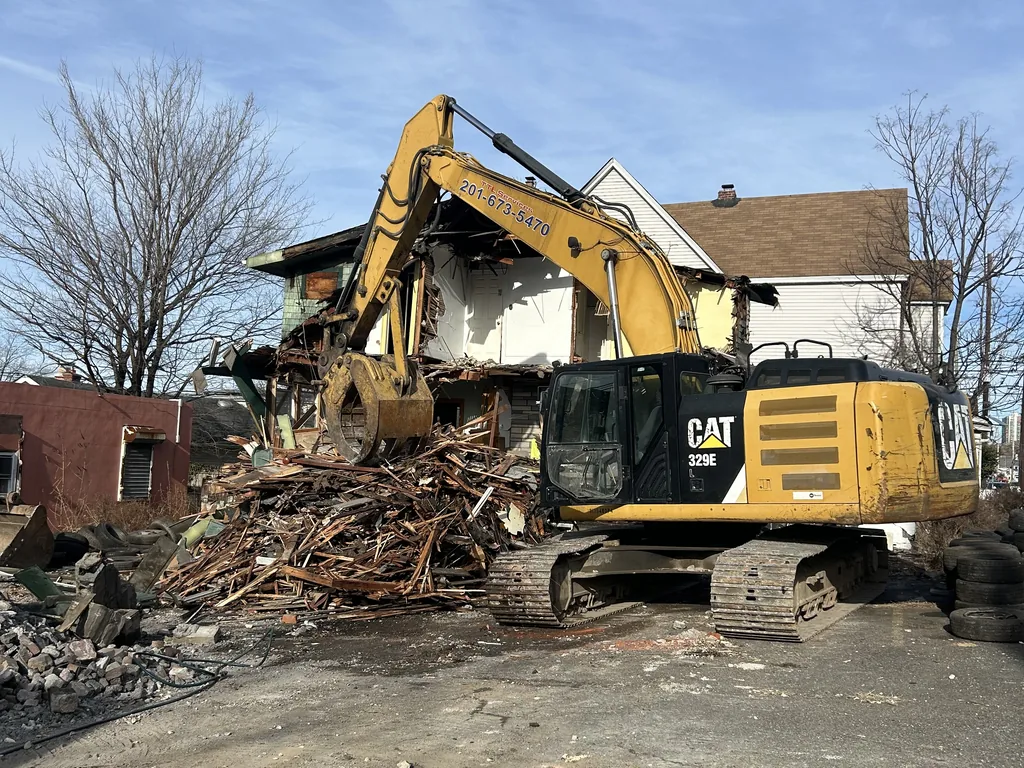 Excavator with a demolition grapple tearing down a two story house, splintered framing piled in front, house demolition