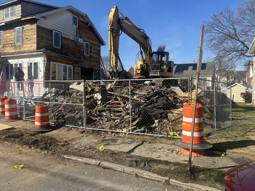 Excavator behind a fenced pile of debris from a house torn down on a residential street, orange barrels at the curb, house demolition