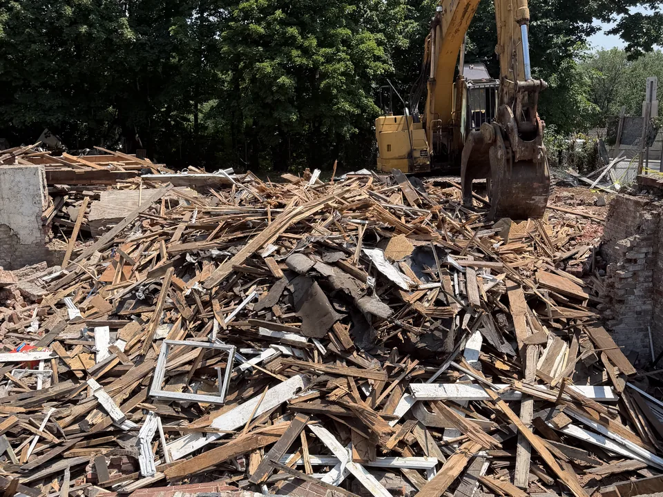Excavator with a grapple behind a large pile of wood framing and brick from a house teardown, house demolition