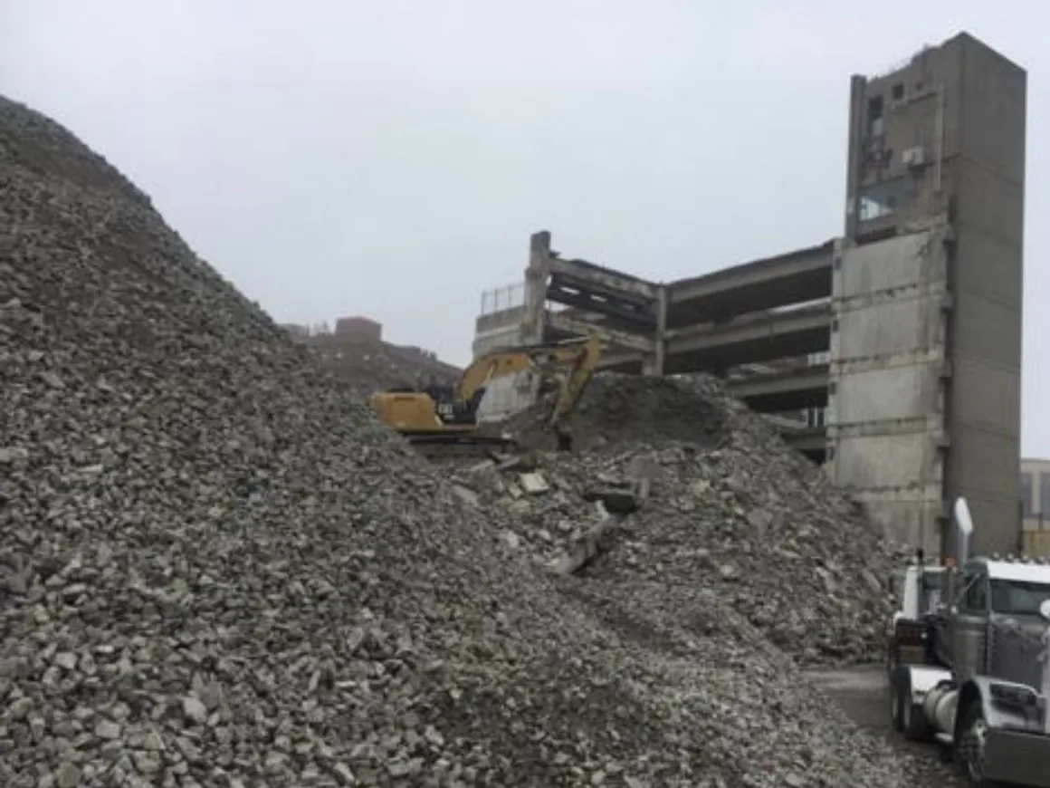 Mountain of broken concrete with an excavator on top beside a partly demolished concrete structure, industrial demolition