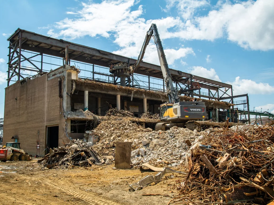 High reach excavator tearing down a concrete and steel mill building, concrete rubble and a heap of scrap steel in front, industrial demolition