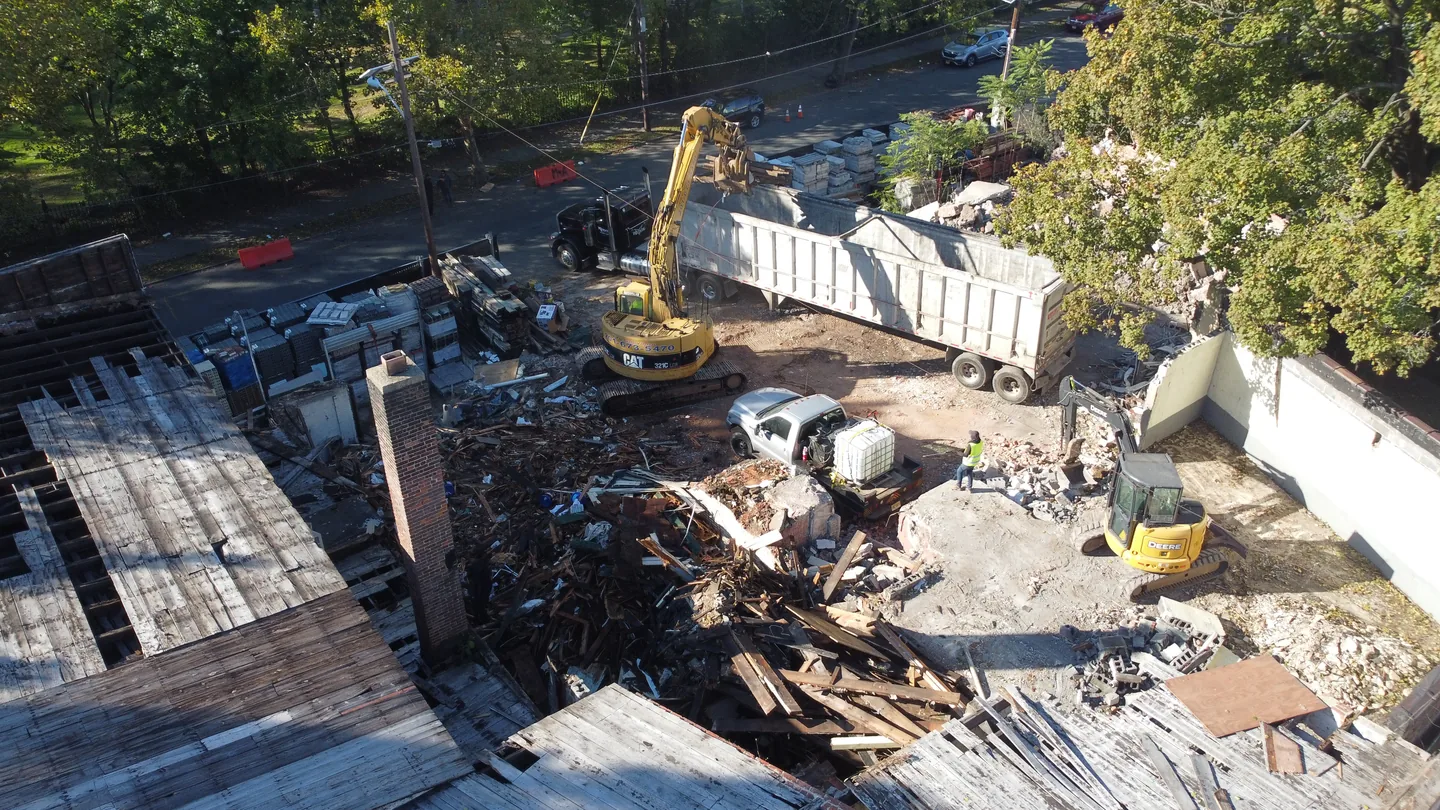 Aerial view of an excavator loading demolition debris into a trailer on a teardown lot by a tree lined street, demolition contractor