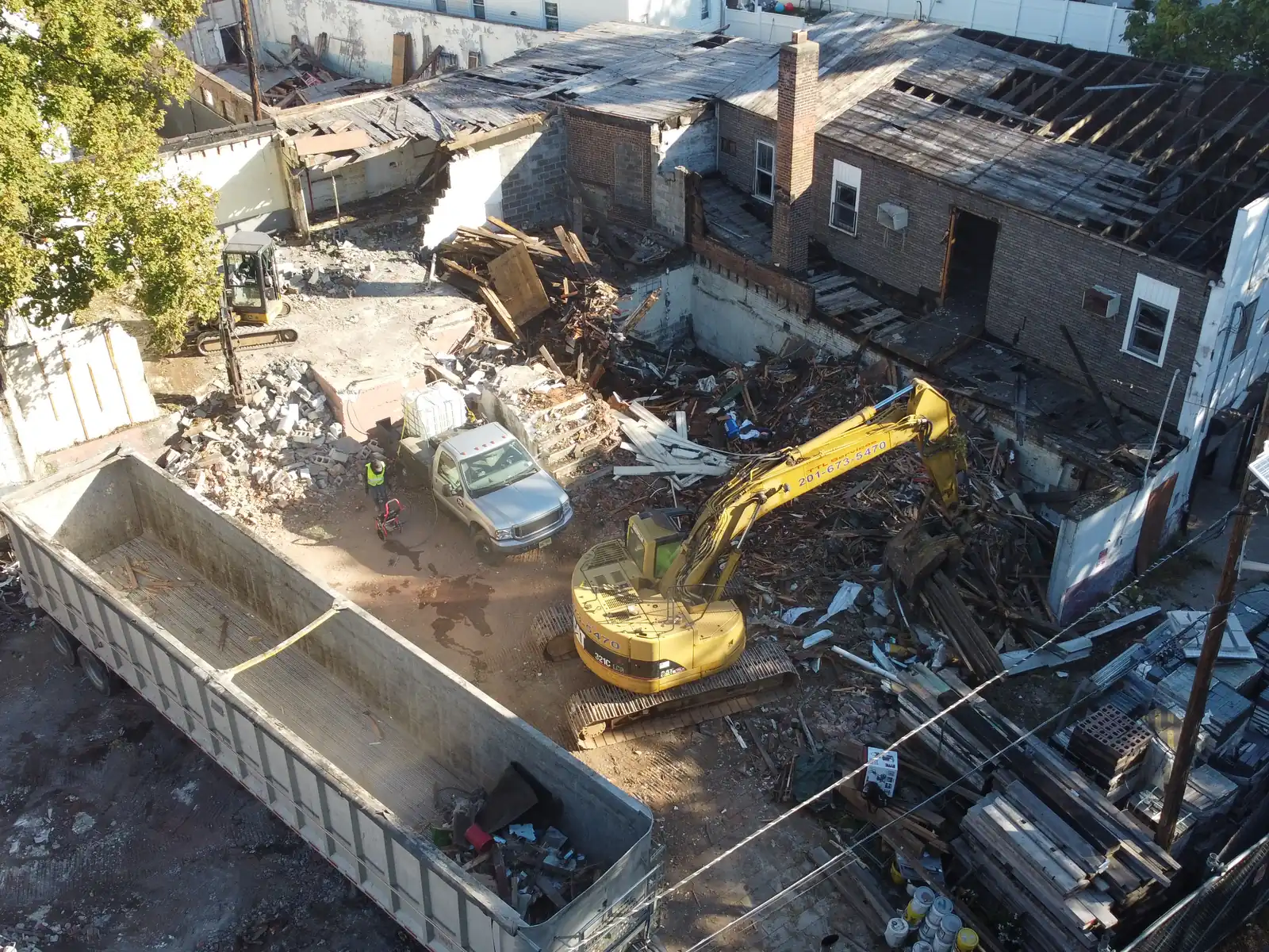 Aerial view of a teardown lot behind a row of buildings, an excavator loading debris into a trailer, a mini excavator beside it
