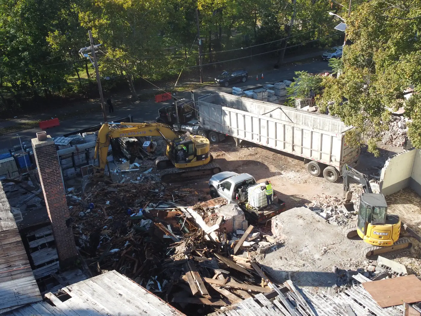 Overhead view of an excavator loading teardown debris into a long trailer parked beside a wall