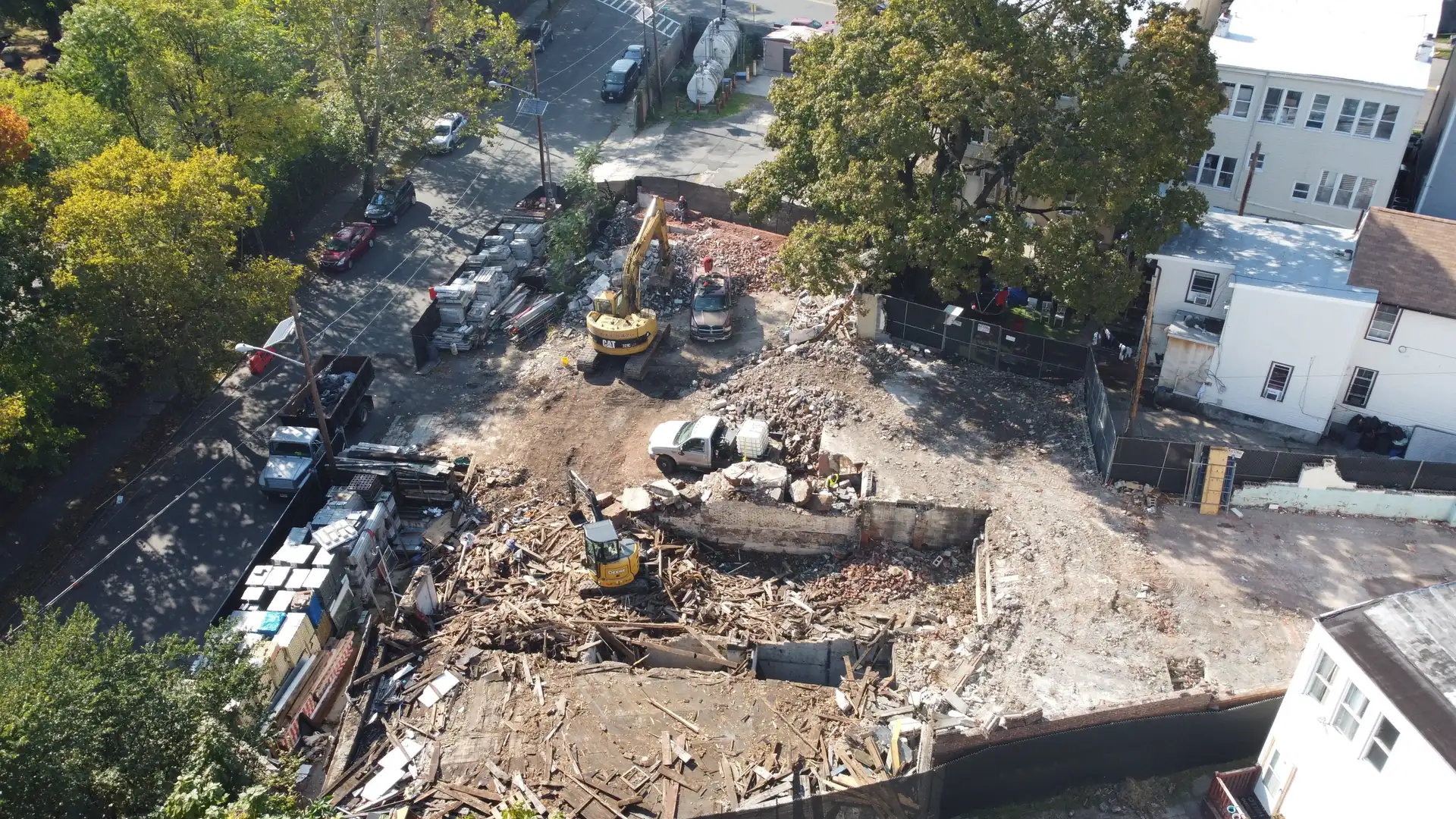 Aerial view of a teardown lot between two buildings, an excavator loading debris into a dump truck at the street, demolition contractor