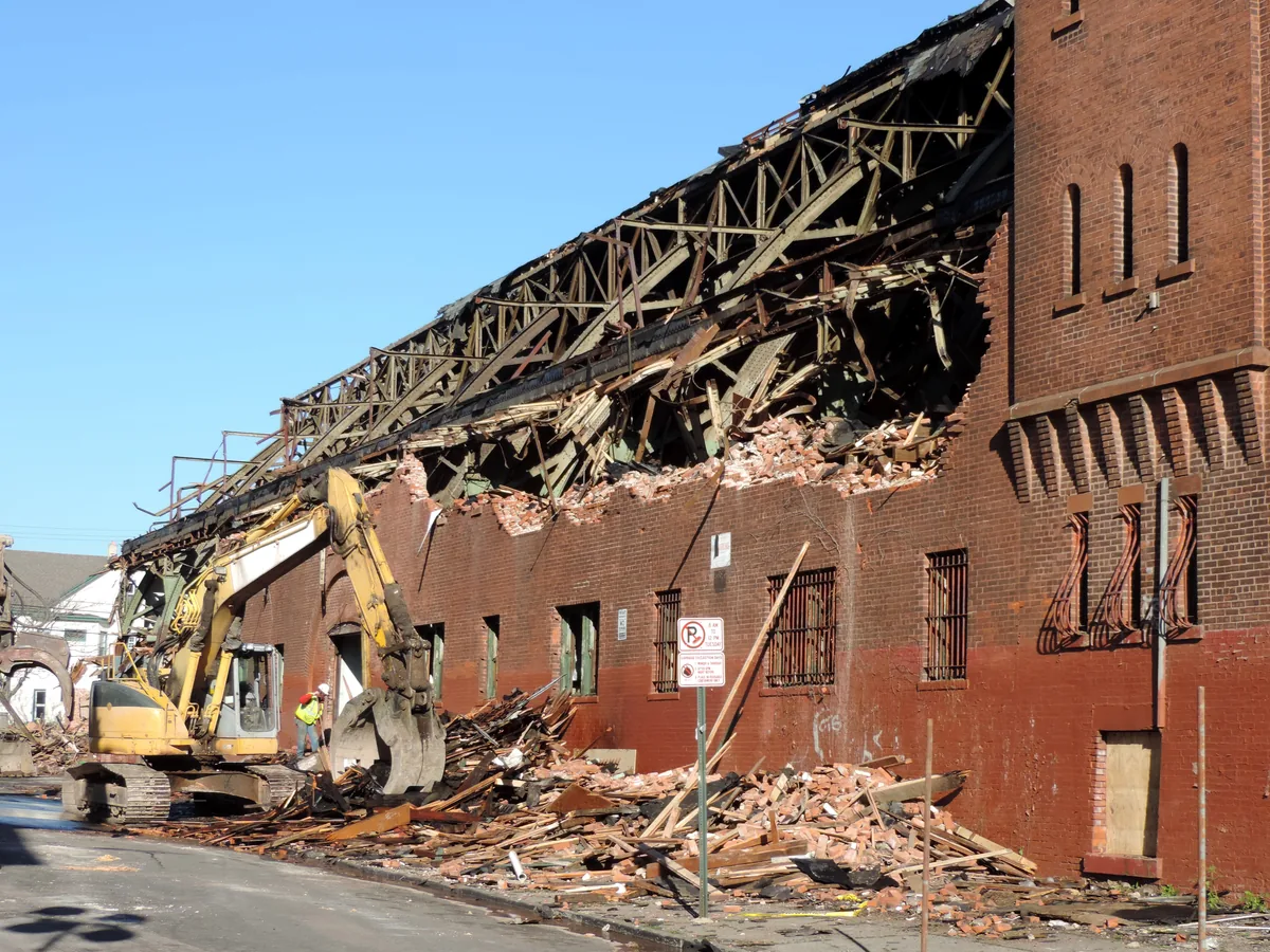 Excavator pulling down the brick wall of a large old building with its steel roof trusses exposed, public works demolition