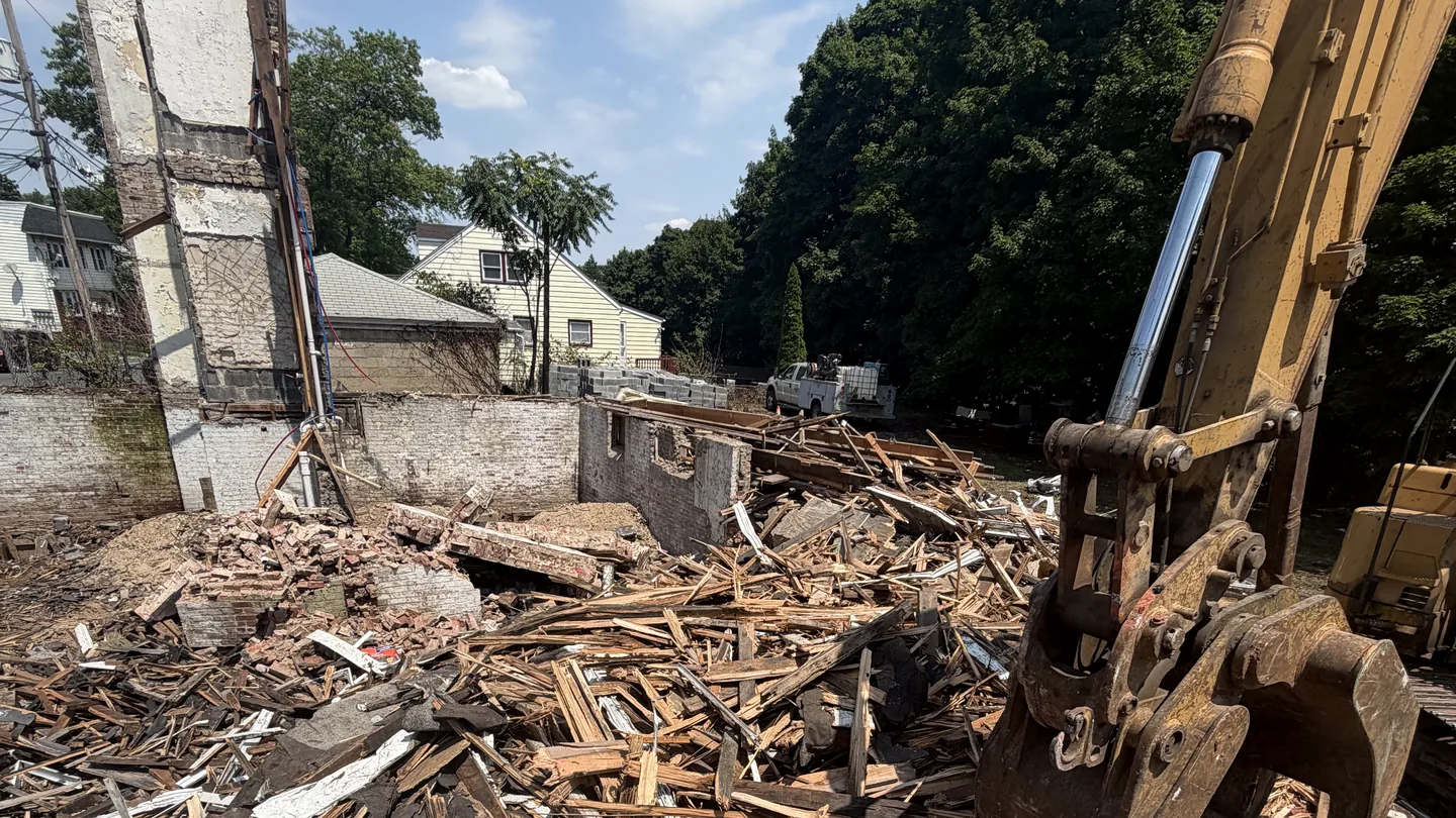 Excavator grapple over the wood debris of a torn down house with its brick stack still standing, demolition contractor service areas