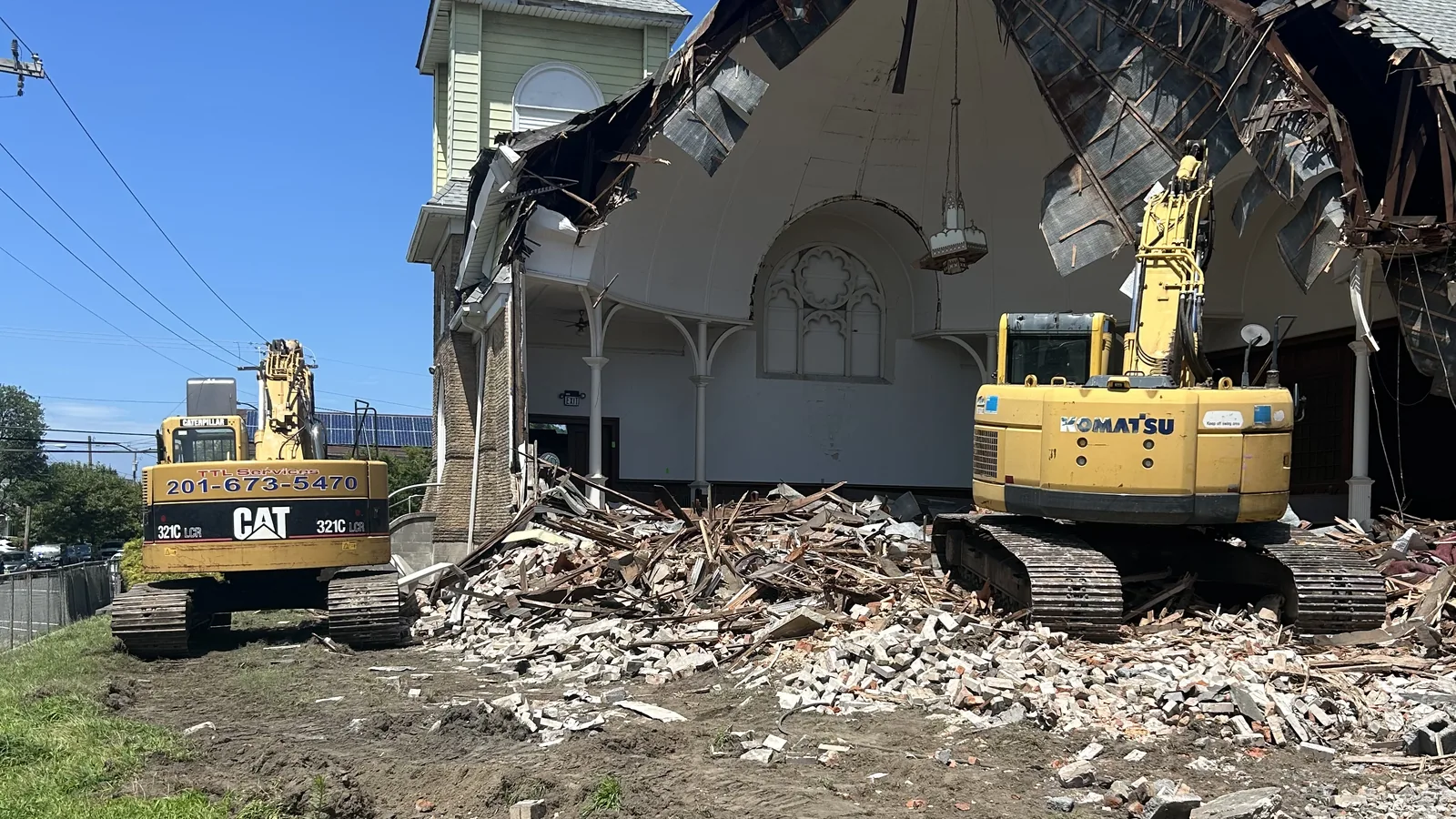 Two excavators taking down a church with its roof torn open below the steeple, a lawn in front, demolition contractor services