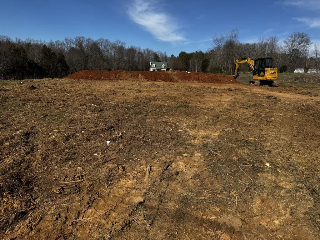 Cleared and grubbed building lot with a compact excavator beside a spoil pile, bare winter woods and a house behind, site preparation