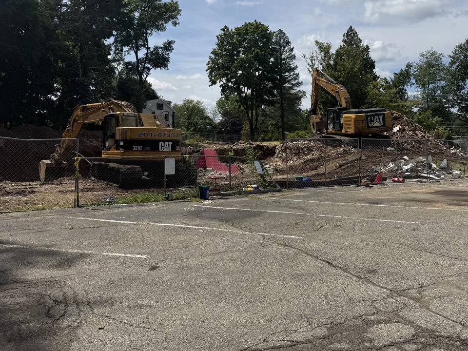 Two excavators on a cleared teardown lot behind a chain link fence, dirt and debris piles, site preparation