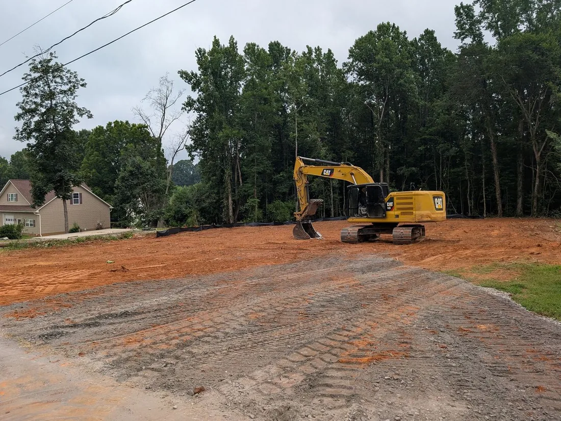 Graded building lot with a tracked excavator, silt fence along the tree line and a gravel construction entrance, site preparation
